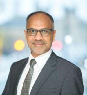 Shailesh is an Asian man wearing black-rimmed glasses. He is wearing a smart black suit with white shirt and smiling at the camera against a blurred background.