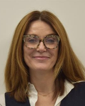 Claire is a white woman with long brown hair, wearing glasses and smiling at the camera against a white background, wearing a white shirt. The image is a headshot.