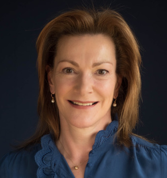 Lady Nicki Hall smiling in a blue shirt with a dark blue backdrop.