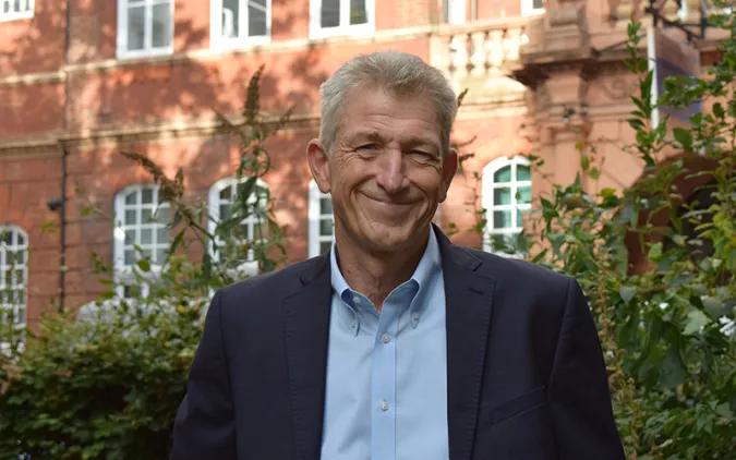 Nick smiling at the camera in a blue shirt and dark suit jacket, against a backdrop of buildings in Queen Square and greenery